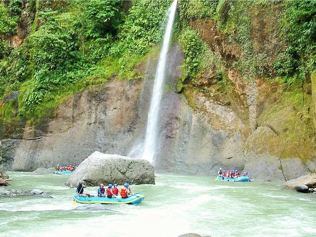 Two rafts navigating a river past a tall, narrow waterfall on a cliff.