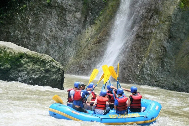 Whitewater rafting group raising paddles together next to a tall waterfall.